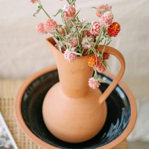 Terracotta style vase with a handle and pink flowers inside sitting on top of a table