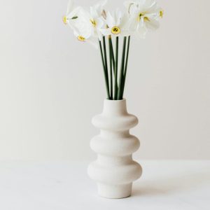 White ceramic vase with a sculptural design holding white flowers with yellow centers, styled on a minimalist white surface