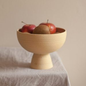 Elevated beige colored bowl filled with red apples and kiwis, sitting on top a table covered with a tan cloth